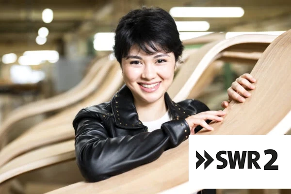 Eine lächelnde Frau mit kurzen schwarzen Haaren und einer schwarzen Lederjacke steht im Innenbereich und hält einen gebogenen Holzgegenstand in der Hand. In der unteren rechten Bildecke ist das SWR2-Logo zu sehen.