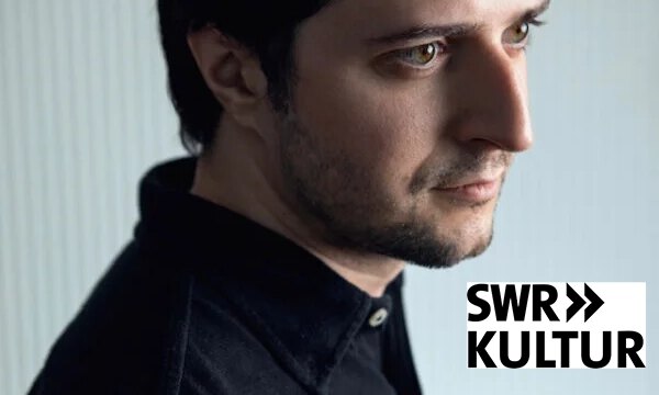 Ein Mann mit kurzem dunklem Haar und gestutztem Bart, der ein dunkles Hemd mit Kragen trägt, blickt nachdenklich zur Seite vor einem hellen, sanft gestreiften Hintergrund.