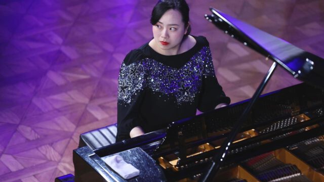 Eine Frau in einem schwarzen Kleid mit silbernen Pailletten spielt auf einem Flügel und blickt nachdenklich auf. Der Boden hat ein geometrisches Holzmuster, und die Beleuchtung wirft einen sanften violetten Farbton auf die Szene.